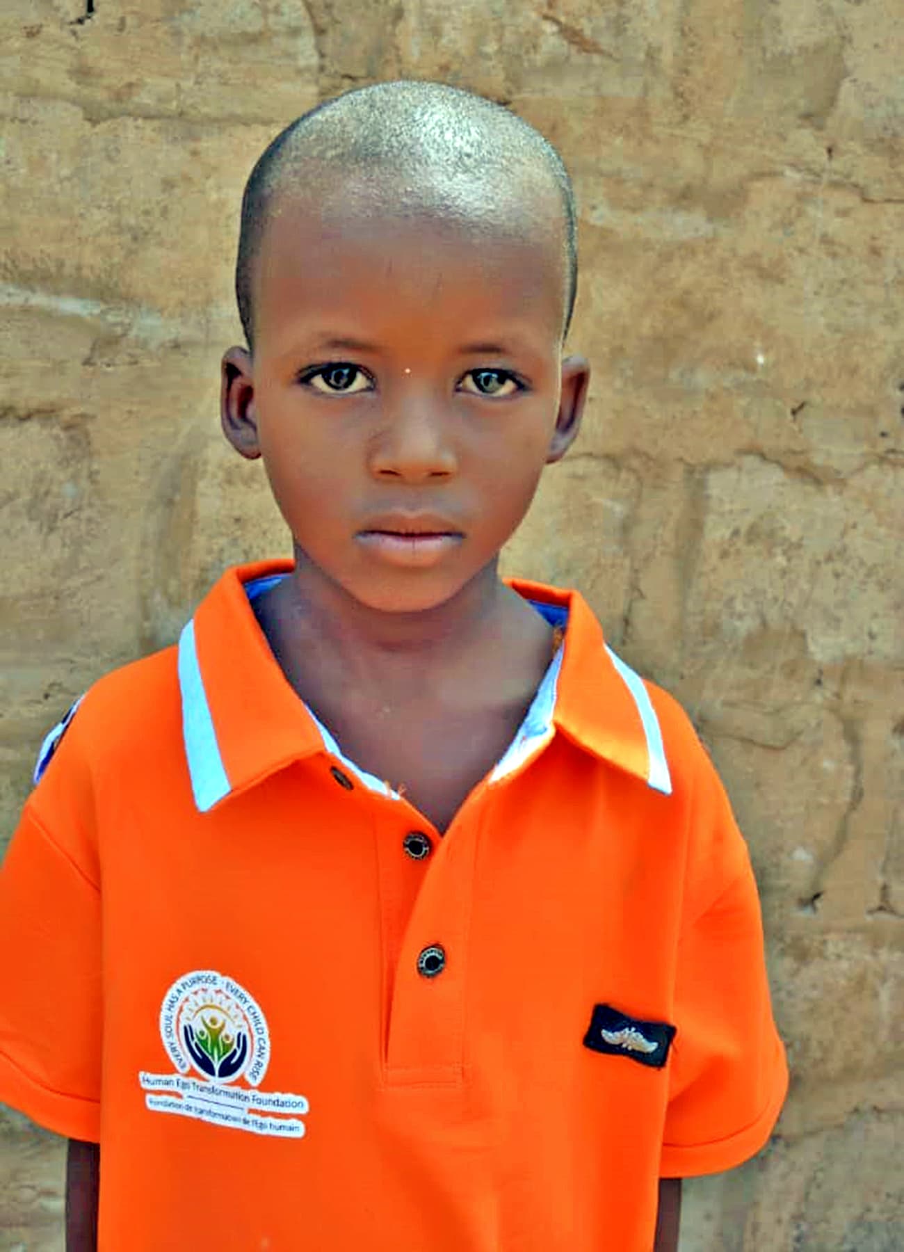 A boy wearing an HET Foundation shirt looks directly at the camera with quiet confidence.